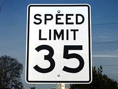 Speed limit sign that reads Speed Limit 35 in front of a blue sky with trees behind it.