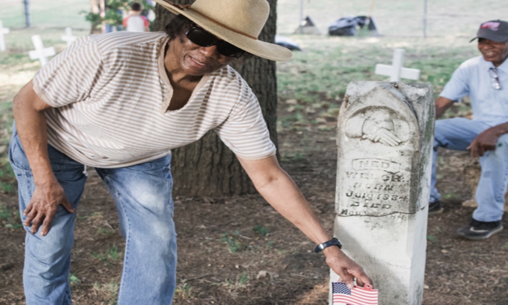 A woman in a hat puts an American flag in front of a gravestone. A man stands behind her smiling.